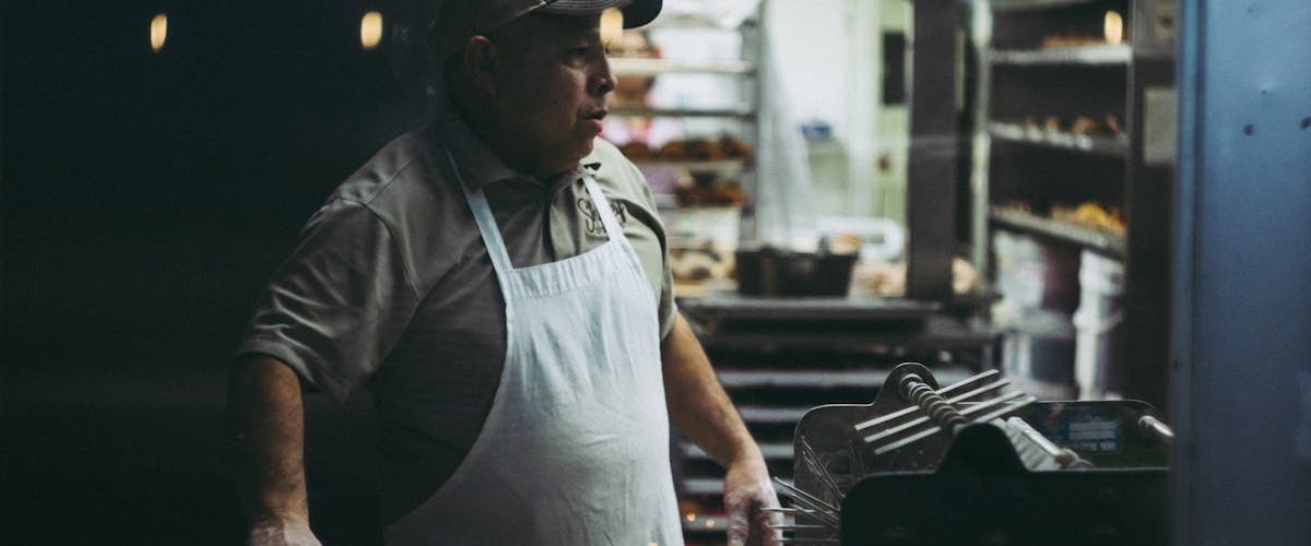 An adult male baker in an apron works in a donut shop kitchen, surrounded by baking equipment.